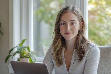 A young woman with long hair sits in front of a window, smiling gently at the camera while using a laptop.