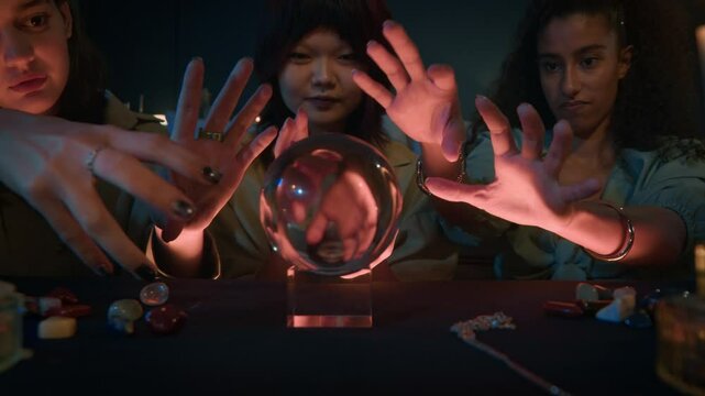 Medium shot of three diverse gen Z women asking crystal ball during fortune telling session at night indoors