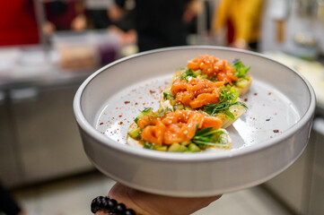 A close-up of beautifully presented dish featuring open-faced toasts topped with fresh salmon and vibrant greens, garnished with red pepper flakes. The background suggests bustling kitchen environment