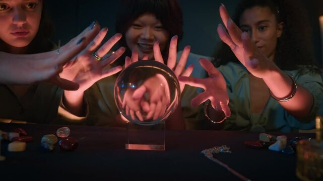 Medium shot of three young multicultural girlfriends predicting their future using crystal ball at night indoors