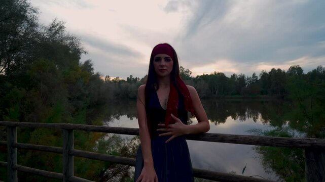 Chica joven realizando movimientos de danza contempor&aacute;nea con un paisaje natural de lago con bosque y el brillante y c&aacute;lido cielo del atardecer dibujando su silueta.