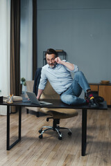 Man is sitting on a desk with a laptop and a cell phone. He is wearing a blue shirt and jeans