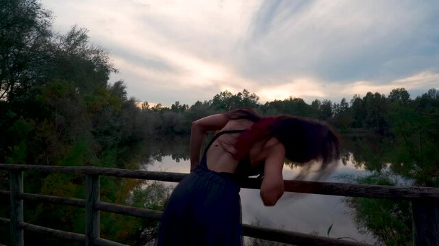 Chica joven realizando movimientos de danza contempor&aacute;nea con un paisaje natural de lago con bosque y el brillante y c&aacute;lido cielo del atardecer dibujando su silueta.