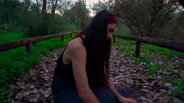 Chica joven sentada en un sendero en el bosque al atardecer, mira hacia abajo y mueve el brazo dibujando algo en el suelo. por mal de amores.