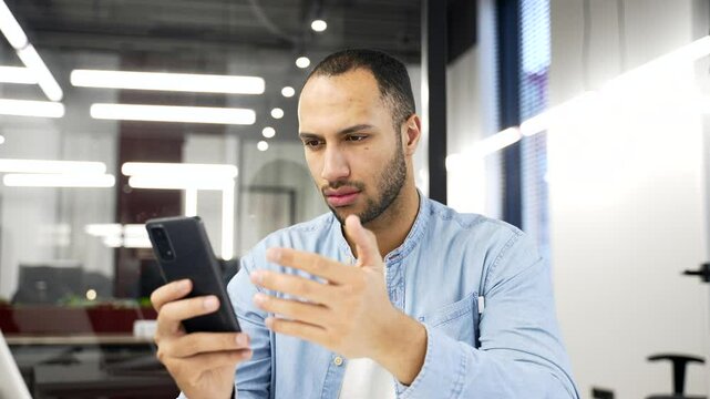 Worried african american businessman having problems with software or service on mobile phone sitting in business office. Frustrated disappointed man complains about bad internet connection
