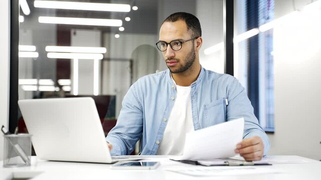Busy thoughtful African American businessman analyzing financial documents checking data on laptop while sitting at a desk at a workplace in a business office. Financier deals with financial report - Powered by Adobe