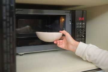 Hand placing ceramic bowl into microwave oven with digital timer display. Close-up kitchen appliance in use. Home cooking and food heating concept.