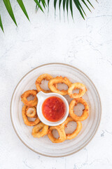 A top-down view of crispy golden calamari rings served on a white plate with a small bowl of red dipping sauce