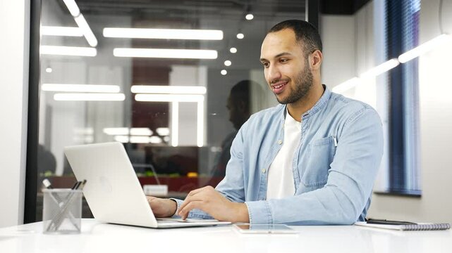 Young african american businessman working on laptop in modern office, smiling while typing. Black man employee using computer application, banking, texting client, chatting online. busy with project