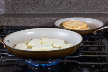 A pan of cheese is cooking on a stove and pitta bread warming up in the background.