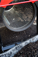 Compost sifter with a wheelbarrow containing mixed and screened soil. Wooden compost bin visible in the background / Concept: Permaculture, Making Compost with motorized compost sifter