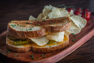 A sandwich with pickles potato chips on a plate with tomatoes on the side. The sandwich is served on a wooden plate.