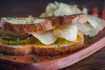 A sandwich with pickles potato chips on a plate with tomatoes on the side. The sandwich is served on a wooden plate.