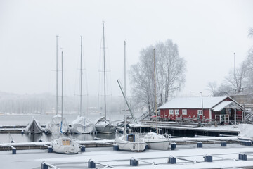White winter scene with small boats in a marina covered by snow with red boat house on the shore on...