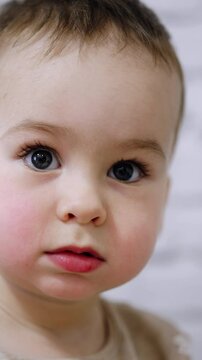 Adorable big-eyed baby boy with mousy hair. Portrait of a calm Caucasian kid focused on something. Close up. Blurred white backdrop. Vertical video