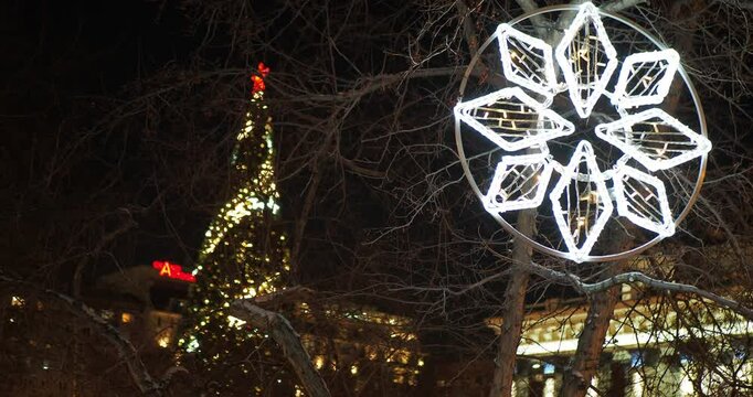 Holiday season, night city. A large snowflake made of lamps glows on a tree against the background of a Christmas tree decorated with burning garlands. The camera is rotating, hand held. Siberia.