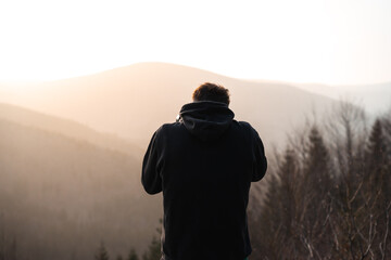 A man dressed in a black hoodie is standing in front of a vast mountain range