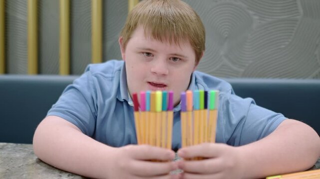 Teenager with Down syndrome having fun with markers. Boy plays game or counts pens after school. Integration of children and adults with disabilities. Lesson with tutor for children with special needs