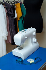 A sewing machine stands on a table with sentimental paper, chalk and pins, against the background of women's clothing and a black mannequin for cutting and sewing.