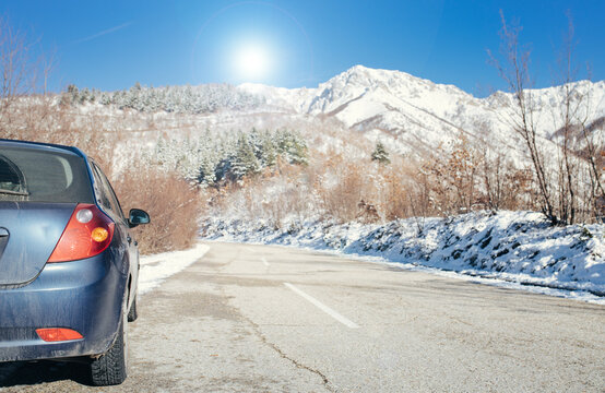 A car parked on a snowy mountain road under a bright blue sky with a sunlit backdrop of snow-covered trees and peaks. A serene and adventurous winter travel scene.