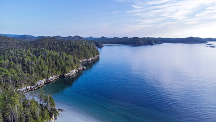 Looking up the coastline of an island in British Columbia Canada. Shore is full of trees growing to the edge of the island. Calm water reflects the mostly clear blue sky 
