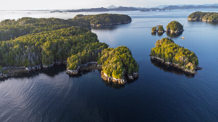 Aerial landscape of forested islands in calm ocean as the yellow light of sunset starts to shine on the trees. In the distance small mountains stretch along the horizon © Matthew