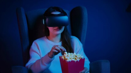 Woman in VR headset watching immersive content with popcorn in dark room. Virtual reality entertainment experience in neon blue lighting. Modern technology lifestyle concept with copy space