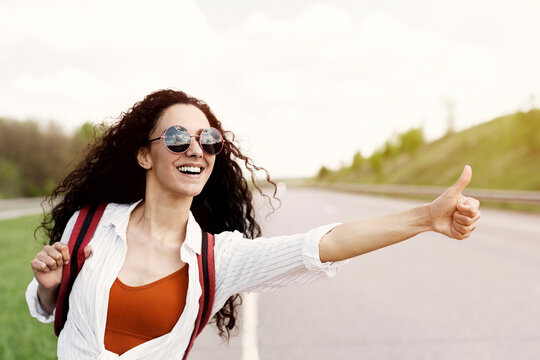 Joyful young lady thumbing her ride, catching car on highway, hitchhiking in countryside, blank space. Lovely millennial female going on summertime adventure, making autostop journey
