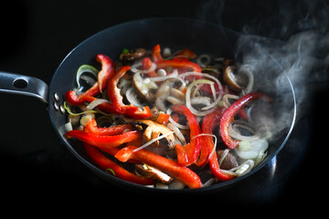 Vibrant red bell pepper slices with mushrooms and onion in a hot steaming frying pan on a black...