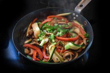 Vegetables in a frying pan such as red bell pepper, zucchini and mushrooms with spring onion garnish on a black ceramic hob, healthy cooking concept, copy space, selected focus