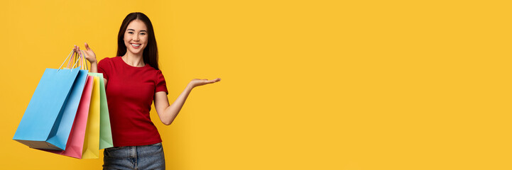 Consumerism, retail. Smiling asian woman with colorful shopping bags showing something invisible product, good on her hand palm, copy space, isolated on yellow studio background