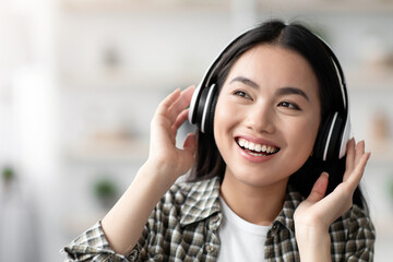 Happy young asian woman in wireless headphones enjoying music while resting on sofa at home, looking at copy space. Portrait of joyful chinese lady enjoying beautiful music, stress relief concept