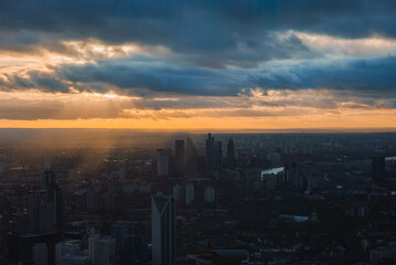 The London skyline is silhouetted against a dramatic sunset sky, featuring the Shard and other skyscrapers. Sun rays pierce clouds, casting a warm glow.