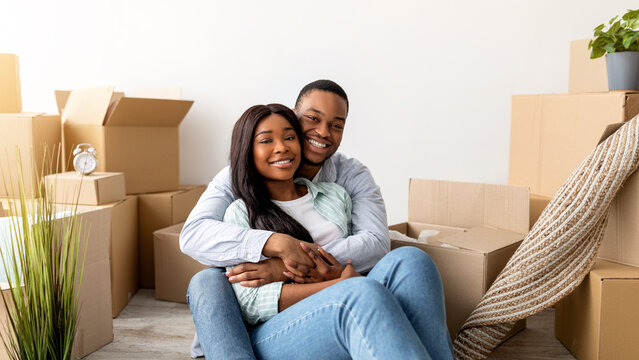 Portrait of loving african american spouses embracing among cardboard boxes, resting on floor after moving to own flat and smiling to camera, copy space