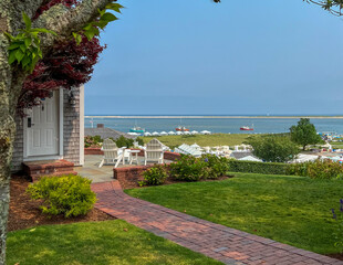 A brick path leads to a cozy patio with white Adirondack chairs overlooking a grassy landscape, vibrant hydrangeas, and a serene harbor with boats under a clear blue sky.