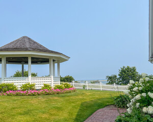 A peaceful garden with a white gazebo surrounded by flowers and a white picket fence, overlooking a serene ocean view under a bright blue sky.