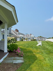 A beautiful coastal lawn with a path leading to Adirondack chairs, colorful flowers, and a white fence, with beach houses in the background and a sunny, peaceful atmosphere.