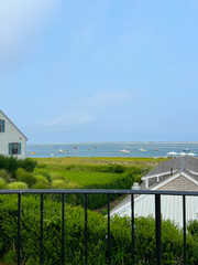 A view of boats on the water with a grassy field and lush greenery in the foreground, framed by a black railing and a charming coastal home on the left under a bright blue sky.