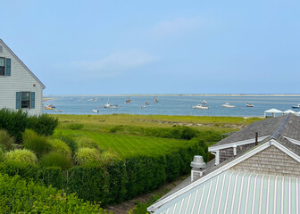 A peaceful coastal view showing boats on the water with a green lawn, shrubs, and rooftops, set against a clear blue sky and grassy shoreline.
