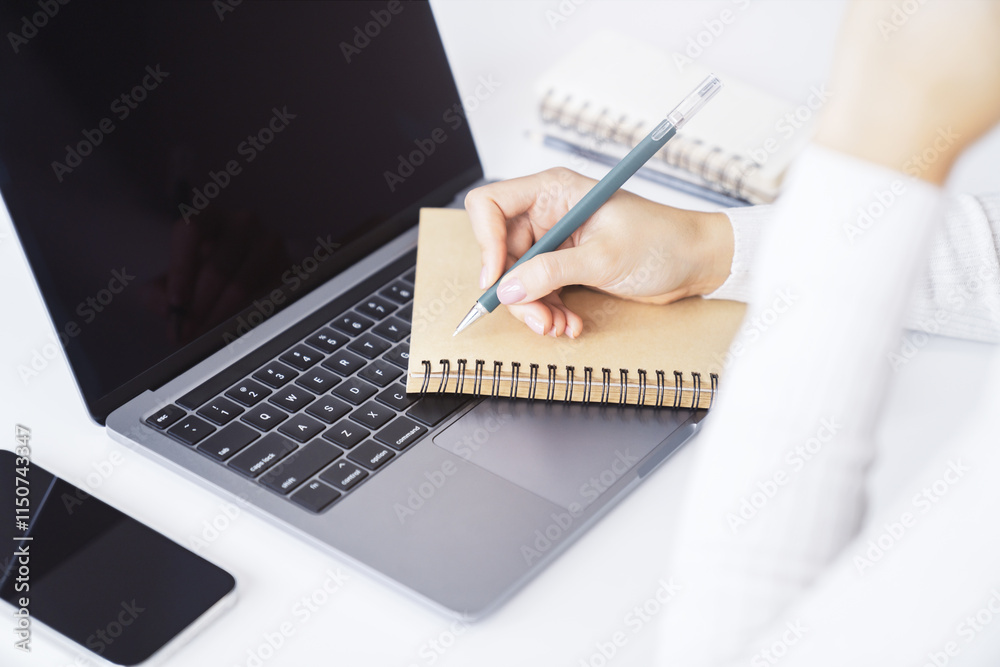 Poster close-up capture of a woman's hand as she writes in a notepad on a sleek laptop, with an out-of-focu