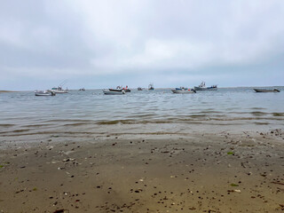 Several boats are anchored near the shore, with some moving through calm waters under a cloudy sky, while the sandy beach in the foreground shows scattered debris and seaweed.
