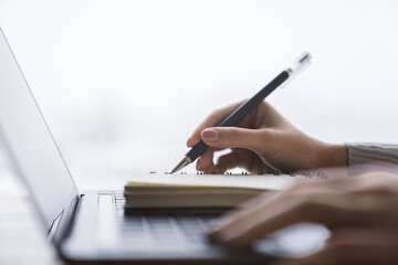 In close view, a woman's hand writing down notes in a notepad on a laptop, against a hazy backdrop