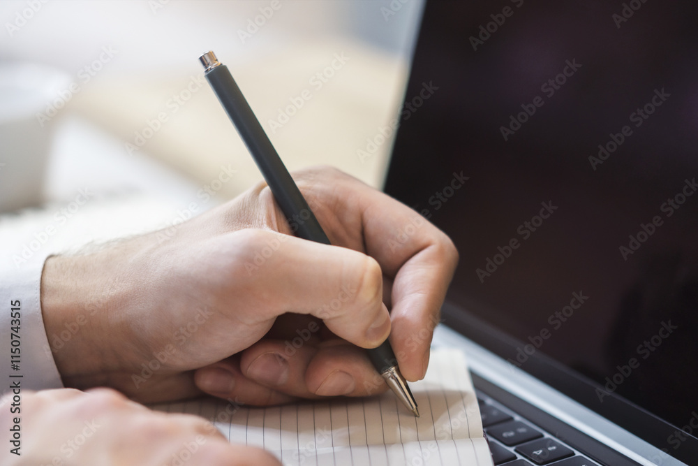 Poster The image focuses on a man's hand scribbling in a notepad on a stylish laptop, set against a blurry scene