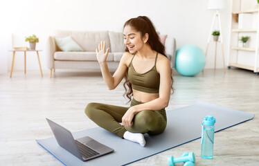 Young fit Indian woman in sportswear sitting on sports mat, greeting her personal trainer online on laptop pc, indoors. Millennial Asian lady ready for web yoga class, communicating on internet