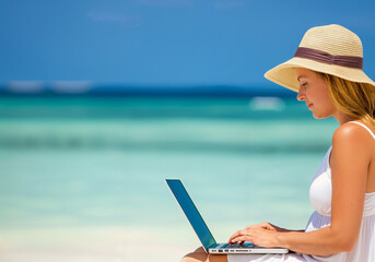 Businesswoman wearing white dress and beach hat working on laptop while enjoying beautiful tropical beach