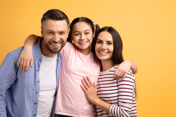 Obraz premium A Caucasian mother, father, and their teenage daughter share a joyful embrace against a bright yellow backdrop, showcasing their strong bond and affection for one another.