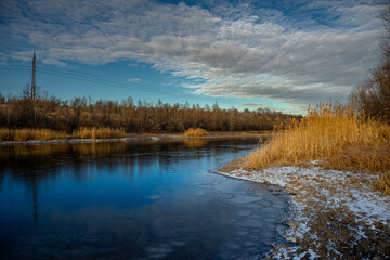 Winter landscape on the pond,frozen pond,blue colors .Landscape with water and ground.Reed on the beach , frozen lake at the morning with sunlights on the sky.Beautiful clouds in the sky,cold and snow