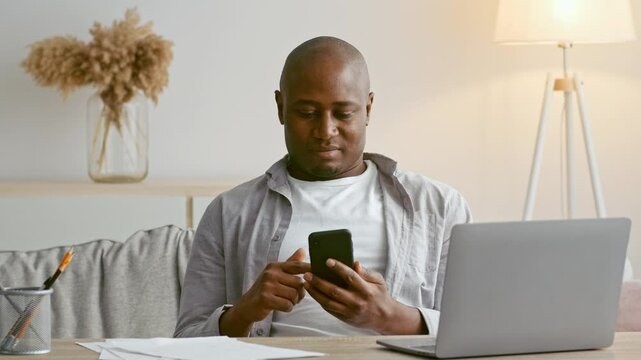 A African American man in his 30s is seated comfortably at home, concentrating on his smartphone while a laptop is open nearby. Papers are scattered around, indicating he is multitasking.