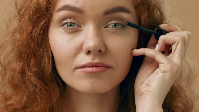 A woman with curly red hair focuses on applying mascara to her eyelashes, highlighting her natural beauty. The setting features neutral tones that enhance the makeup application.