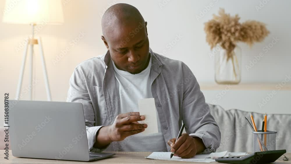Poster a man in his 30s is focused on writing notes while reviewing a document in his comfortable home offi